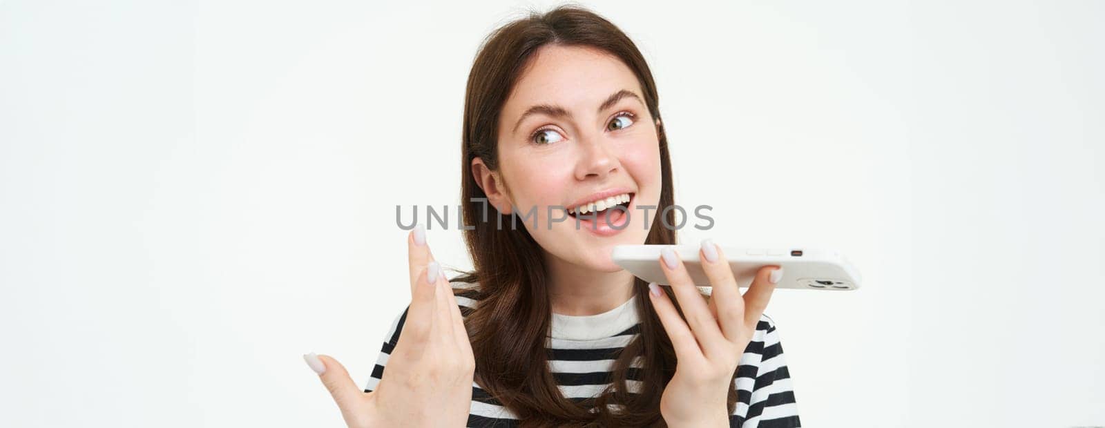 Image of cute brunette woman talking into speakerphone, holding mobile phone near mouth, records her voice, sends a voicemessage, using online translator app, white background.