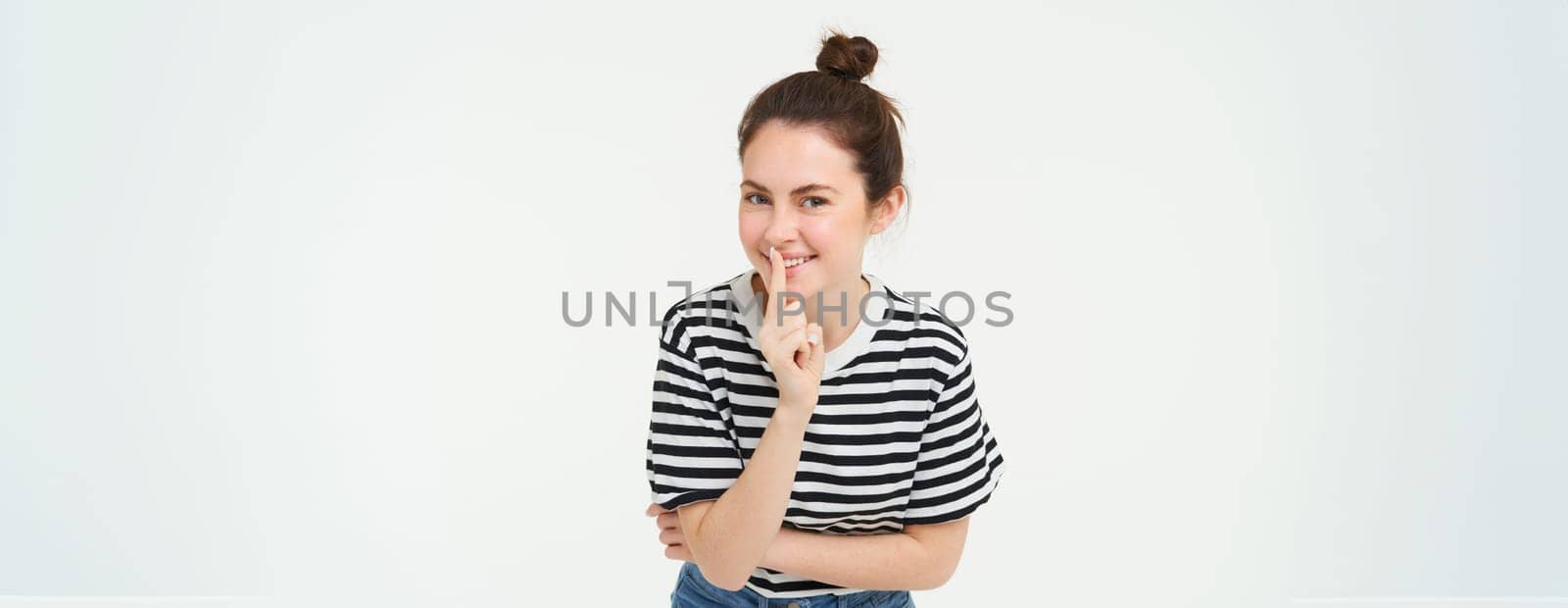 Beautiful, coquettish girl, holds finger on lips, promise to keep secret, shh gesture, hush sign, stands over white background.