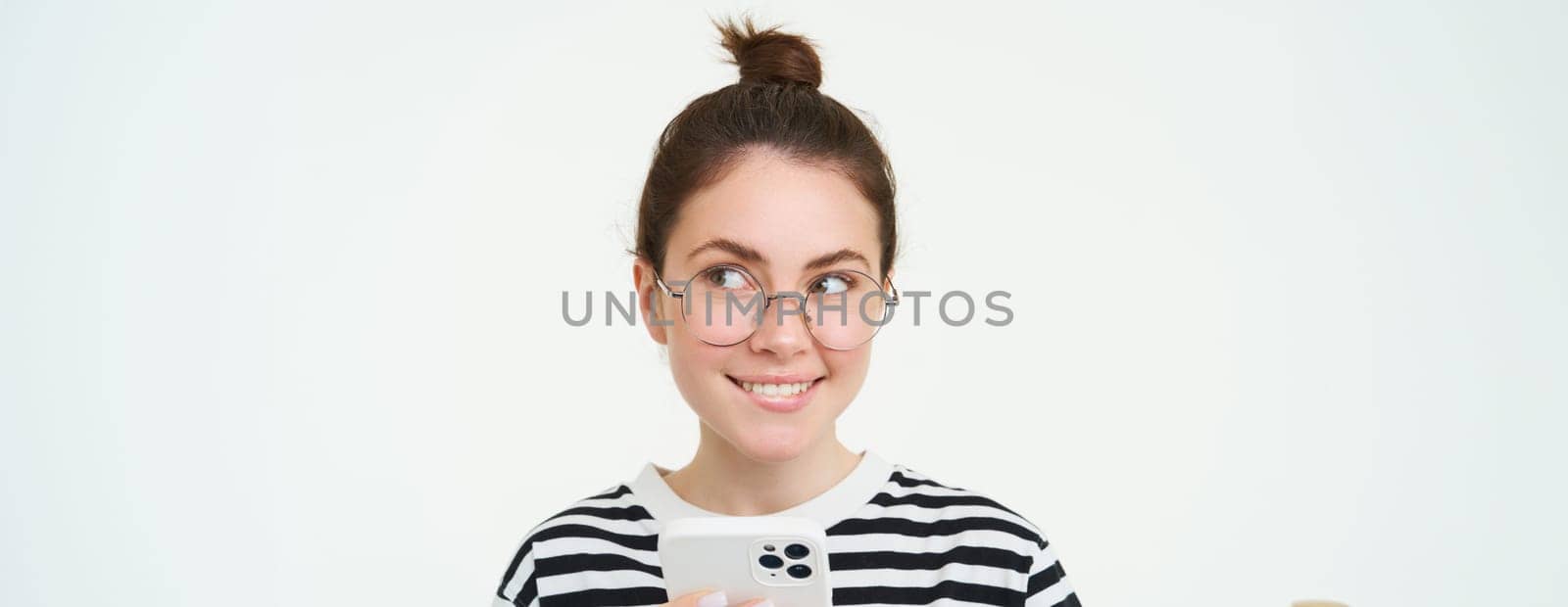 Image of young woman in glasses, using her mobile phone, standing with smartphone and smiling, standing over white background.