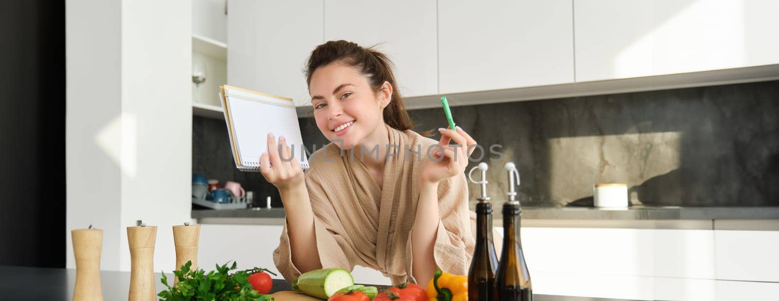 Portrait of good-looking, smiling girl with grocery list, holding notebook and reading recipe, cooking breakfast, making meal with vegetables, preparing salad by Benzoix
