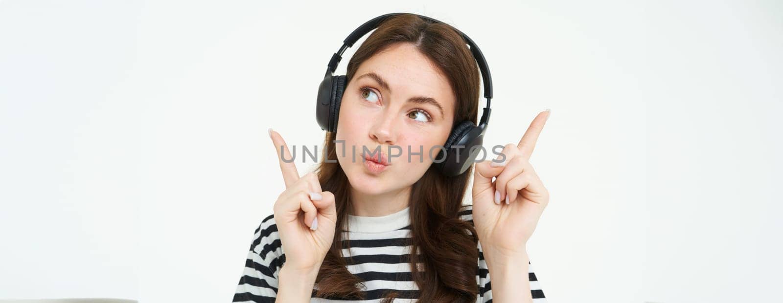 Portrait of young brunette woman in headphones, listening music in headphones, pointing fingers up, showing banner, advertisement, isolated over white background by Benzoix