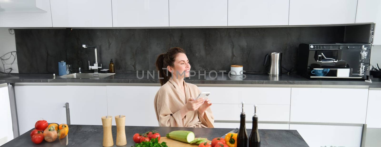 Portrait of woman in bathrobe sitting in kitchen with smartphone, cooking dinner, watching recipe on social media, video tutorial how to prepare meal by Benzoix