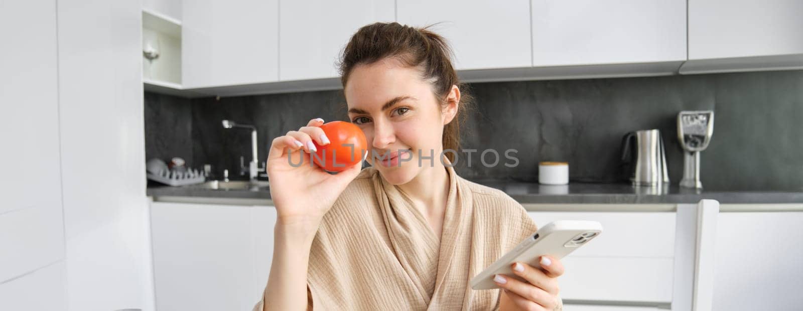 Close up portrait of beautiful smiling woman, holding fresh tomato, sitting in kitchen with smartphone, orders vegetables online, using application to buy groceries, using mobile phone by Benzoix