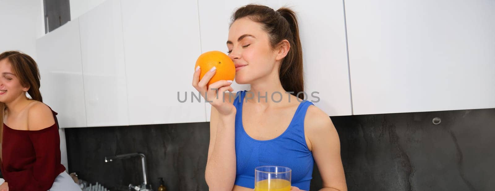 Portrait of fitness instructor, smiling woman, enjoys smell of fresh fruit, drinking orange juice and looking happy, standing in kitchen in activewear by Benzoix