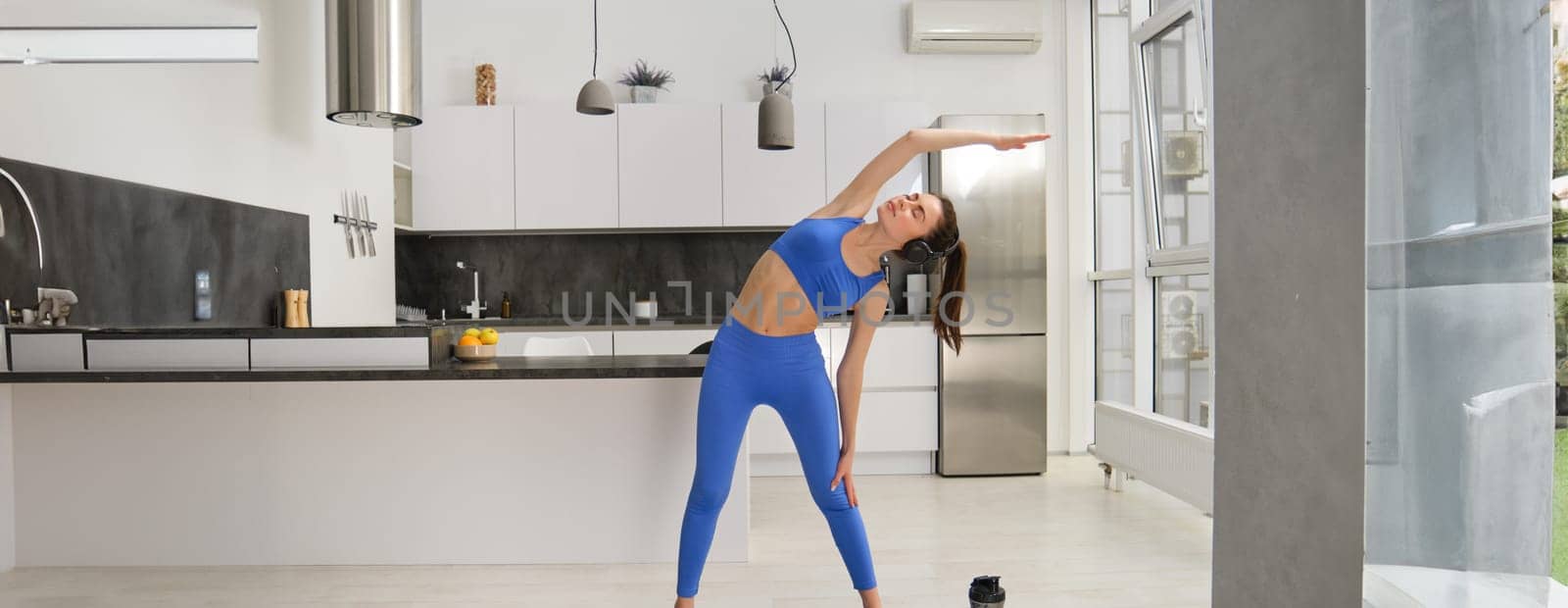 Image of young woman doing fitness exercises at home, stretching her body, bending sideways, doing aerobics workout in living room, standing on rubber mat.