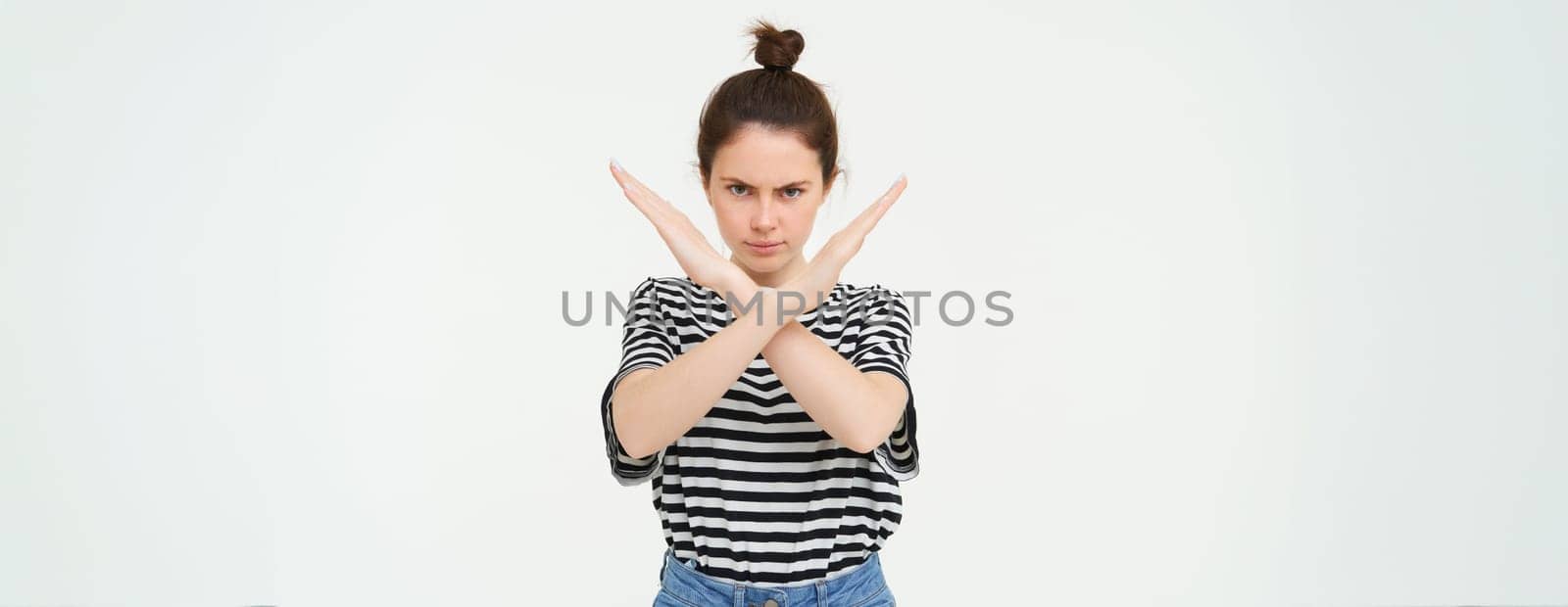Portrait of young serious woman, shows cross, stop gesture, prohibit something, expresses her disapproval, stands over white background by Benzoix