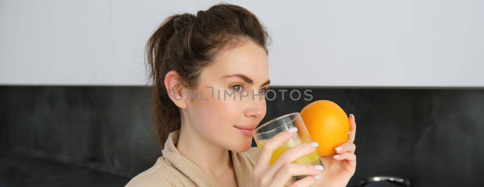 young brunette woman drinking orange juice in white modern kitchen and smiling, girl holding glass of fresh juice, healthy food and drink concept.