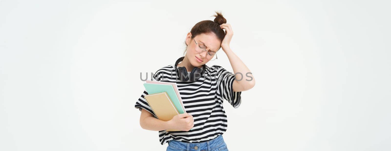 Disappointed young woman, looking tired or upset, wearing glasses, holding study material, documents and notebooks, stands over white background by Benzoix