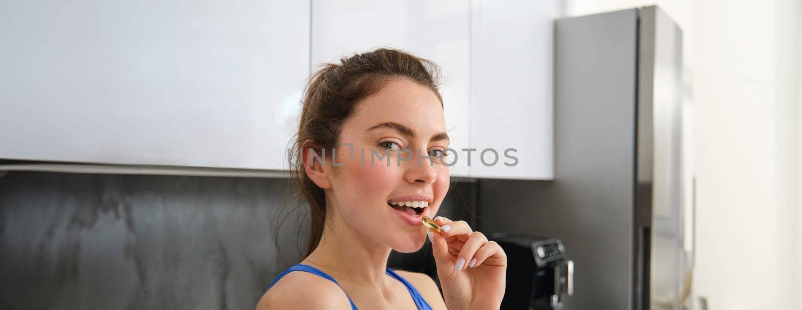 Close up portrait of smiling sportswoman, fitness girl taking vitamins, fish oil pill, using dietary supplements for strong health and fit body, standing in kitchen.