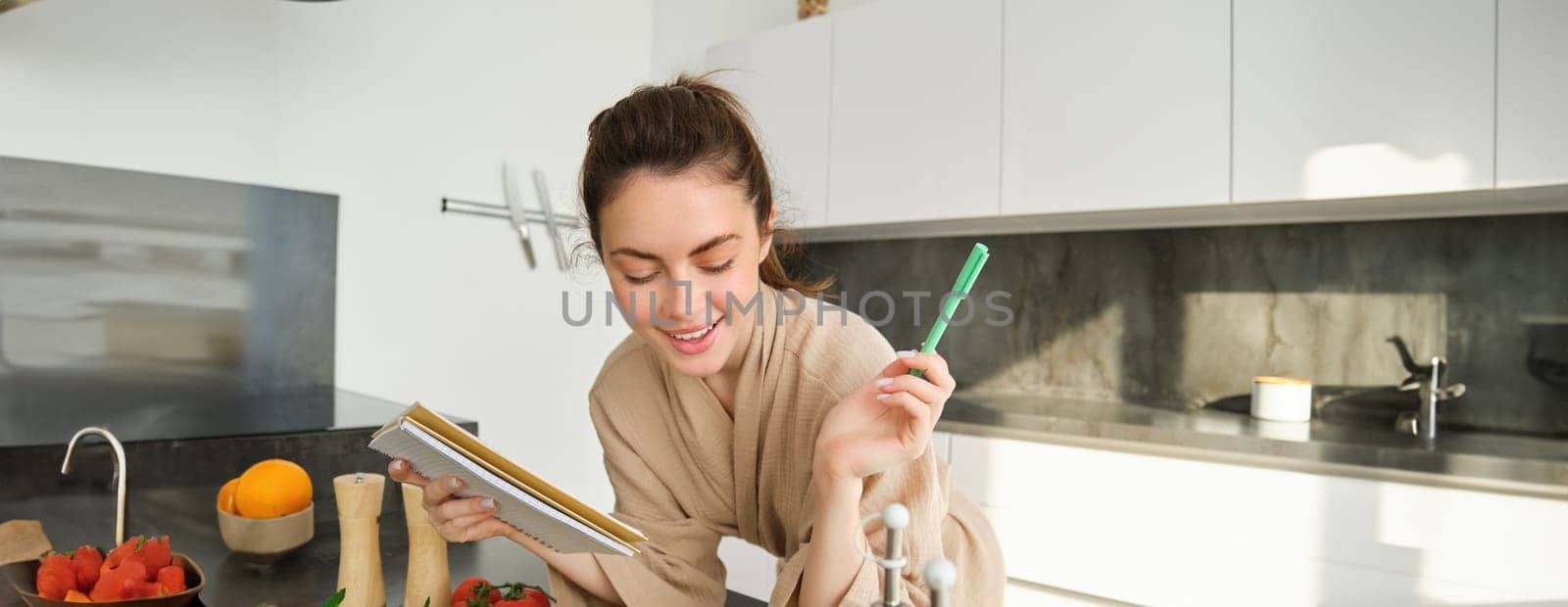 Portrait of woman checking grocery list, looking at vegetables, holding notebook, reading recipe while cooking meal in the kitchen, chopping tomatoes and zucchini.