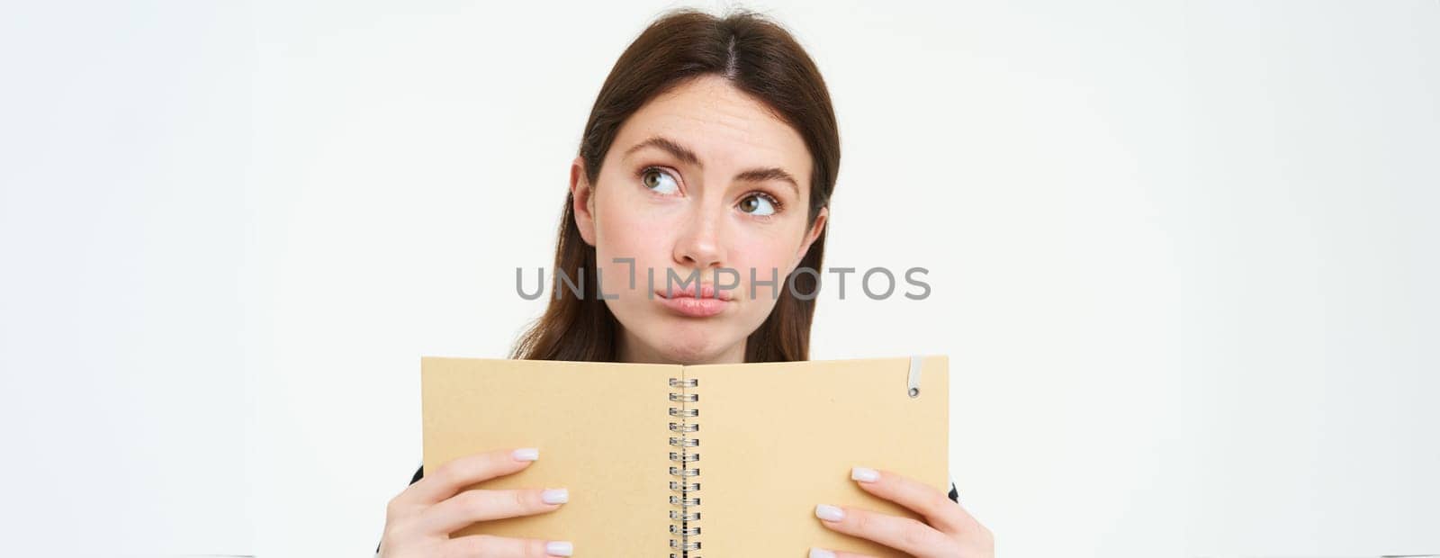 Portrait of thinking girl holding a notebook. Young woman with thoughtful face, holding weekly planner, checks her schedule, writes notes in her memo book, stands over white background.