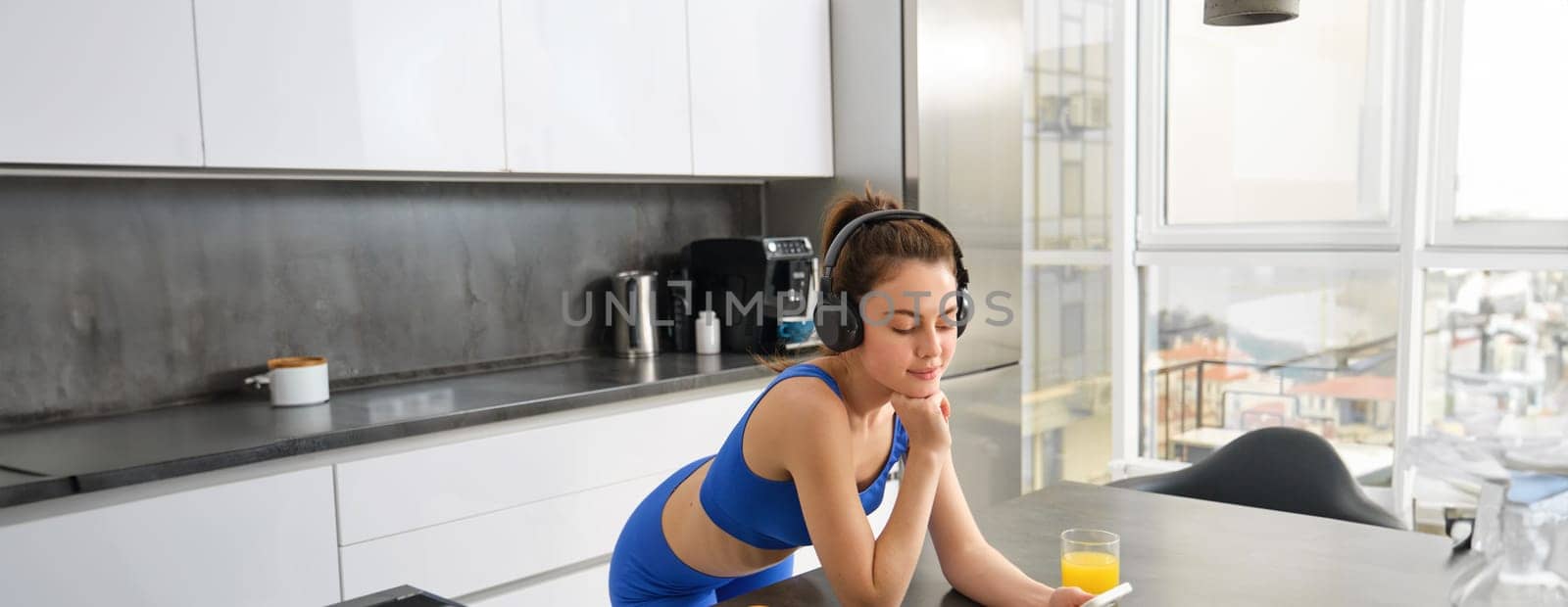Portrait of young modern woman, beautiful girl in fitness clothes, standing in kitchen, watching yoga video class on smartphone, listening in wireless headphones, drinking juice.