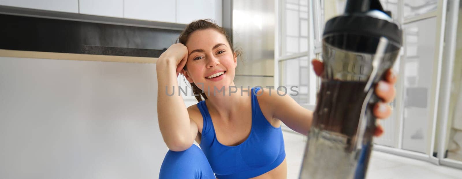 Close up portrait of happy, healthy young woman, give you sports water bottle, stays hydrated during fitness session, workout indoors at home.