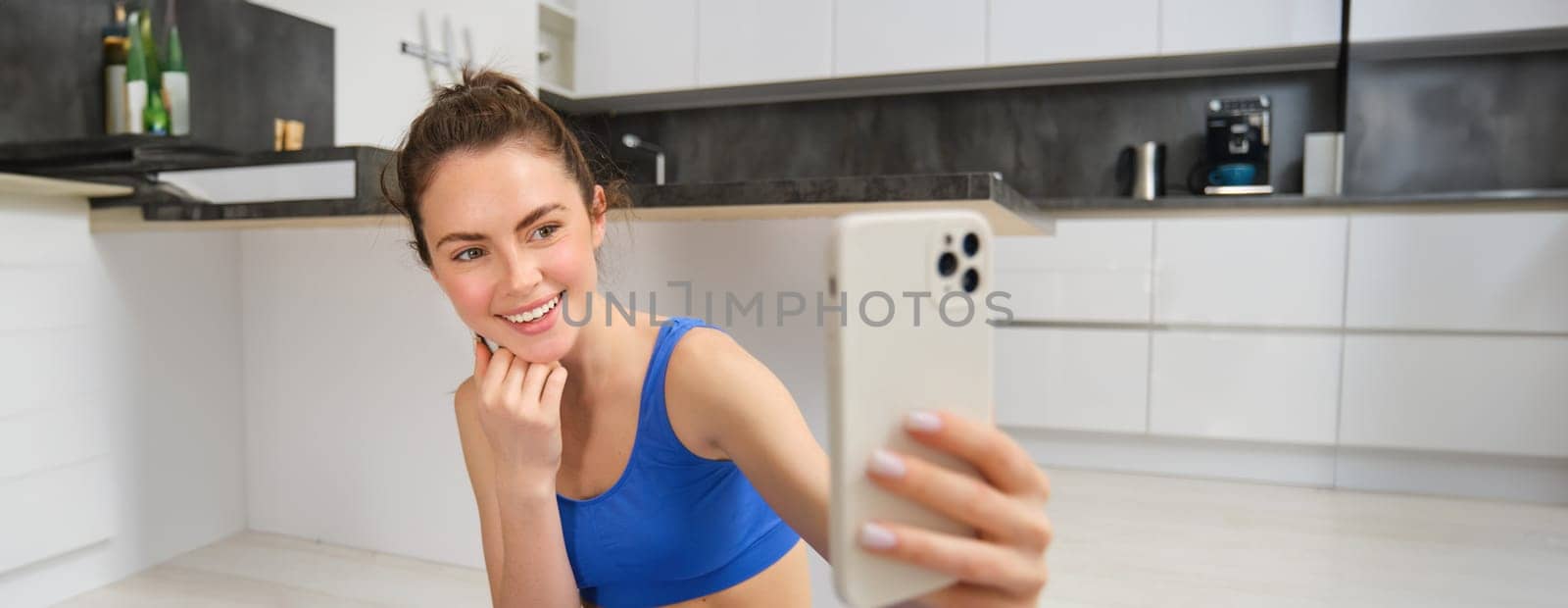 Woman doing workout training from home and taking selfie on smartphone camera, posing for photo inside her house, sits on rubber yoga mat in blue leggings and sportsbra.