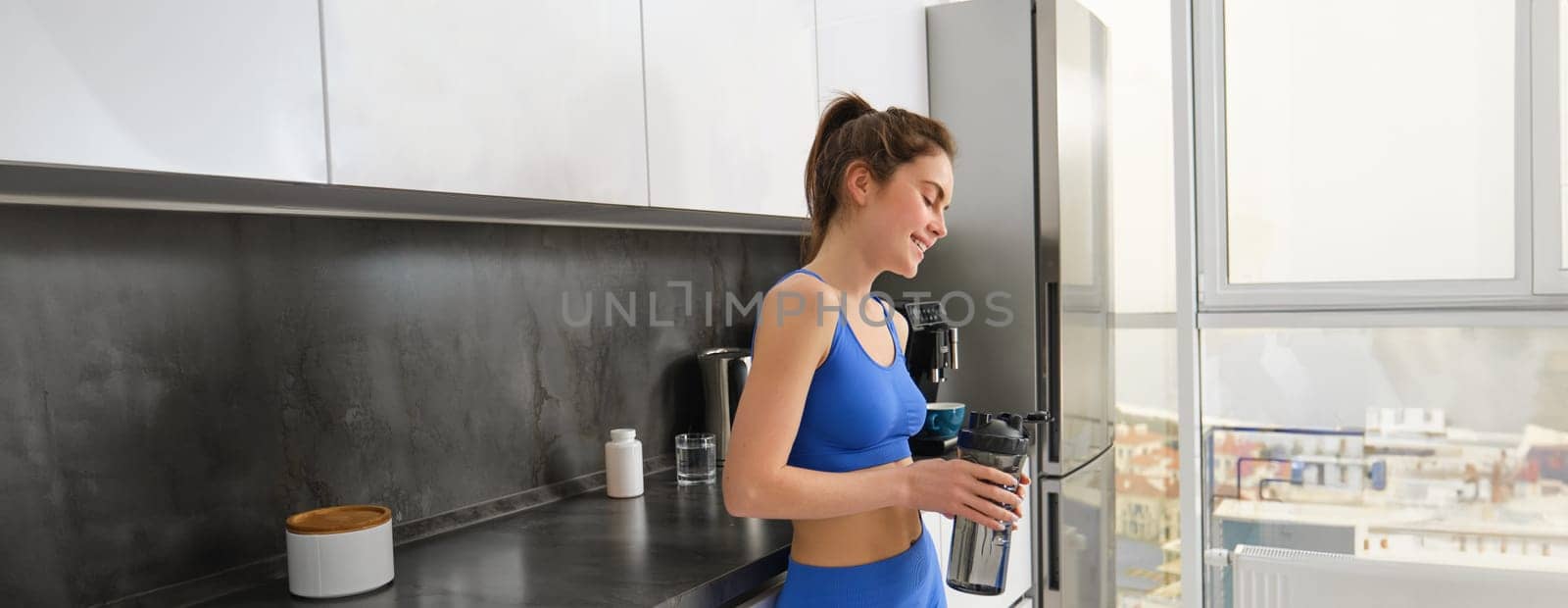 Image of young fitness instructor, woman in sportsbra and leggings, holding water bottle, drinking after workout, standing in kitchen.