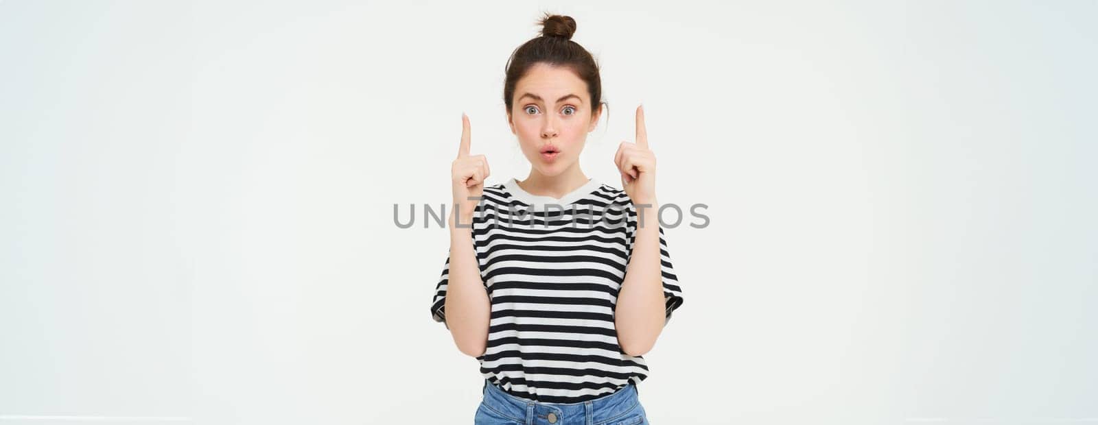 Emotion of amazement. Young woman with impressed face, says wow, points fingers up, shows promo banner on top, recommends product, white background.