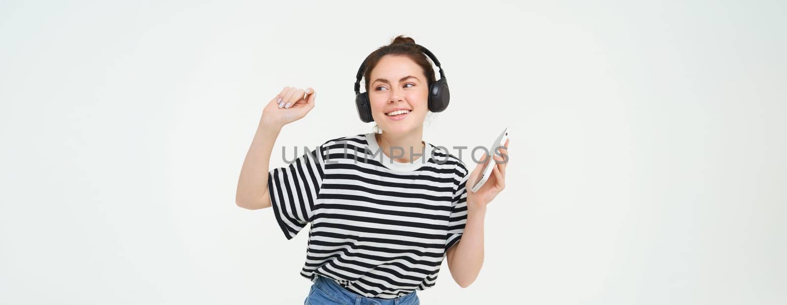 Young woman with smartphone listening to music, dancing to her favourite song in headphones, posing against white background.