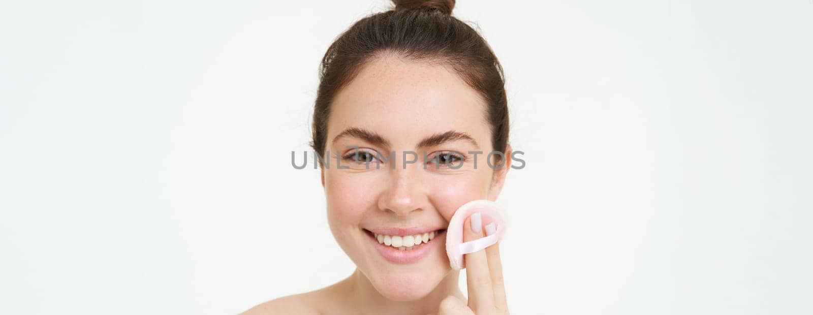 Portrait of smiling happy woman without blemishes, using cosmetic pad to clean her face, using skincare routine treatment, facial toner, takes off makeup with cleanser, stands over white background.