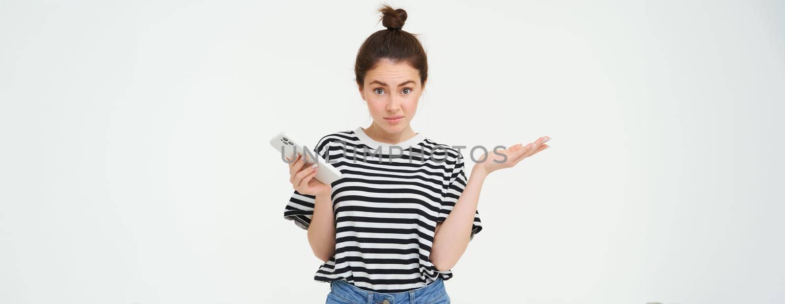 Portrait of shocked girl shrugs shoulders, looks confused reaction to something she saw on mobile phone, stands over white background.