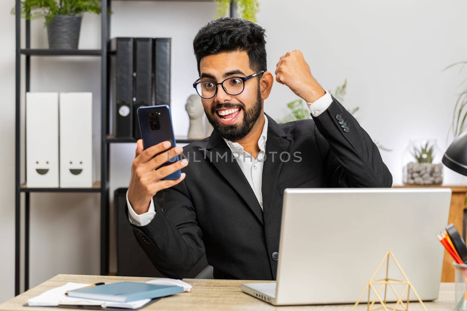 Happy Arabian businessman freelancer use mobile smartphone celebrating success victory winning lottery jackpot goal achievement, play game, good positive news, triumph working at home office workplace