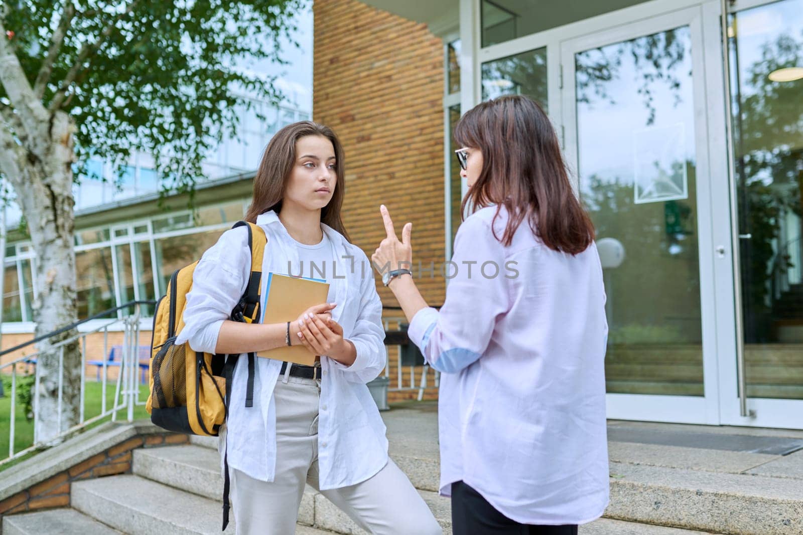 Teenage girl high school student with backpack talking to female teacher, mentor, coach, standing outdoors on steps of educational building. Adolescence, education, knowledge, communication
