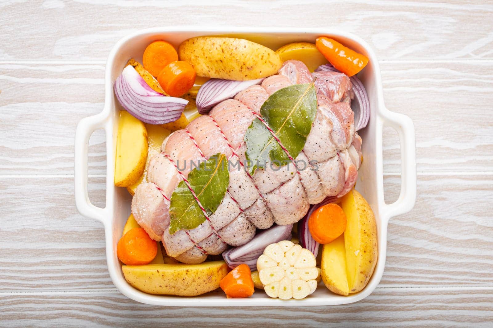 Raw wrapped rolled sliced pork in white casserole dish with potatoes, vegetables and herbs on rustic white wooden background top view. Pork roll with vegetables ready to be prepared.