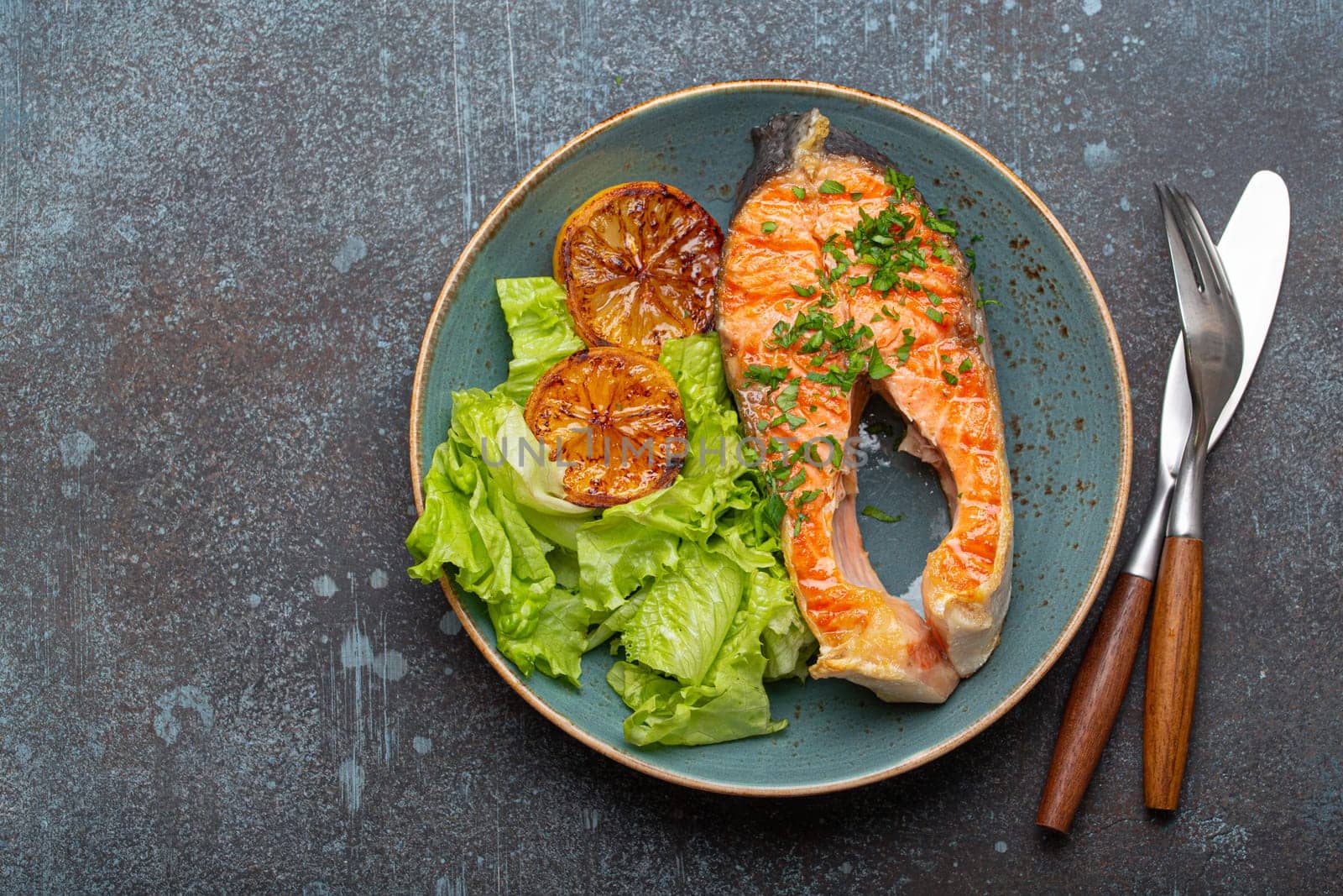 Grilled fish salmon steak and green salad with lemon on ceramic plate on rustic blue stone background top view, balanced diet or healthy nutrition meal with salmon and veggies, space for text.