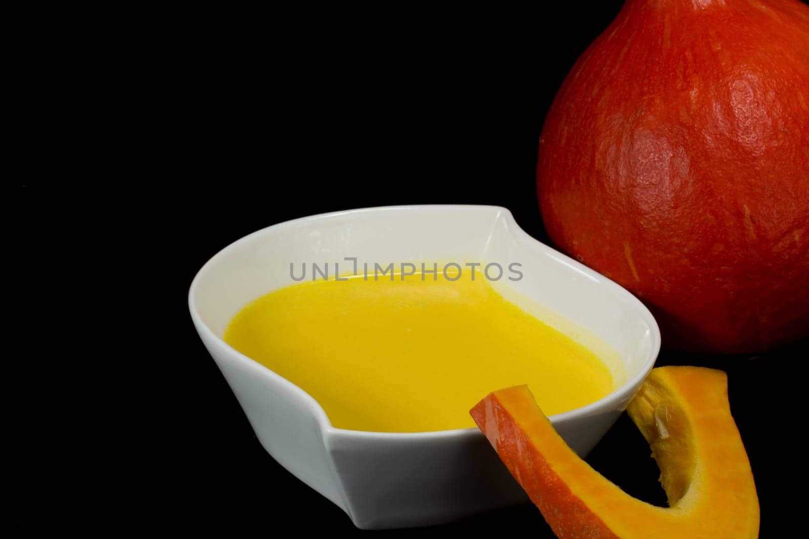 White bowl of pumpkin soup with cream. One piece of pumpkin is leaning on a bowl. Red pumpkin is in the background. All isolated on black background.