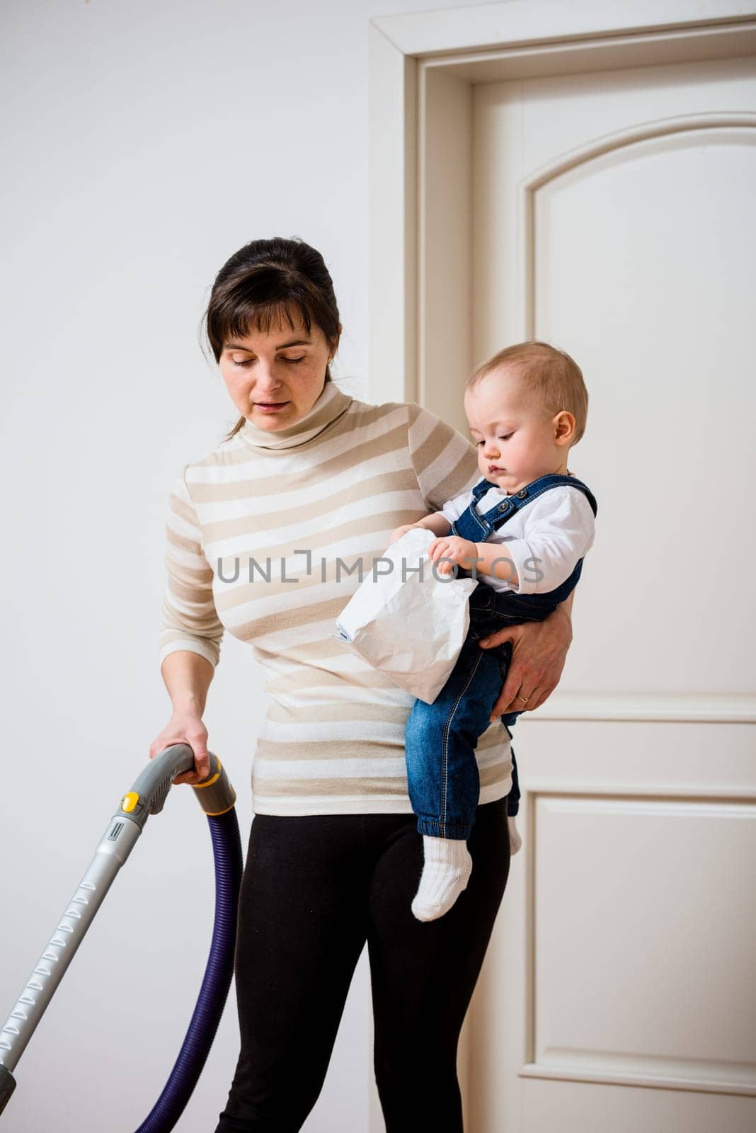 Cleaning up the room - mother with vacuum cleaner holding her baby on hands