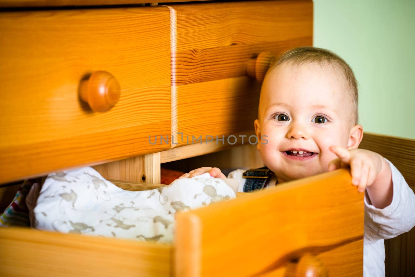 Baby opening drawer with clothes on wooden furniture at home