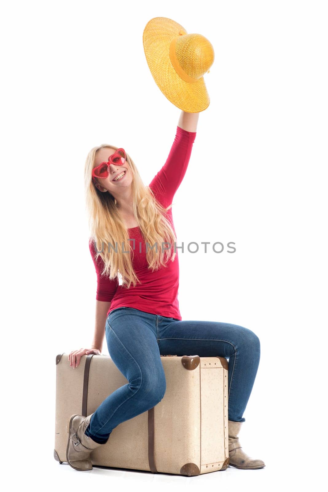 a woman sits on a suitcase and waves with her summer hat
