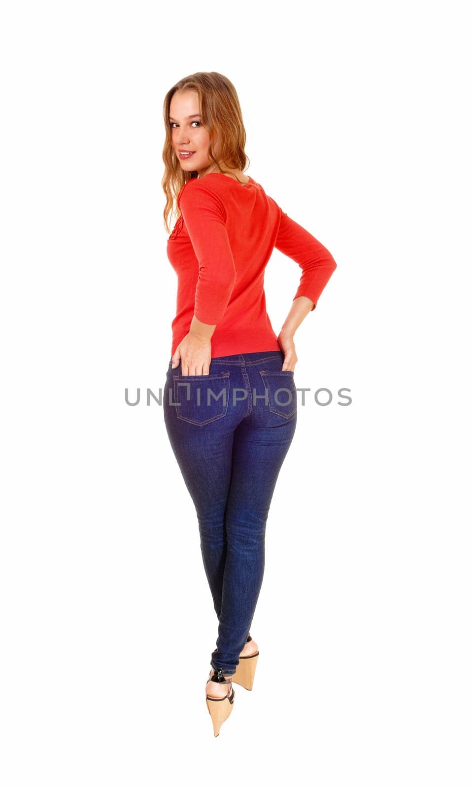 A beautiful blond woman standing from the back in jeans and a red.sweater, looking over her shoulder, isolated on white background..