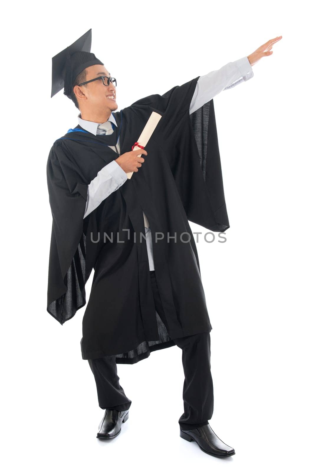 Full length happy pan Asian university student in graduation gown, standing isolated on white background.