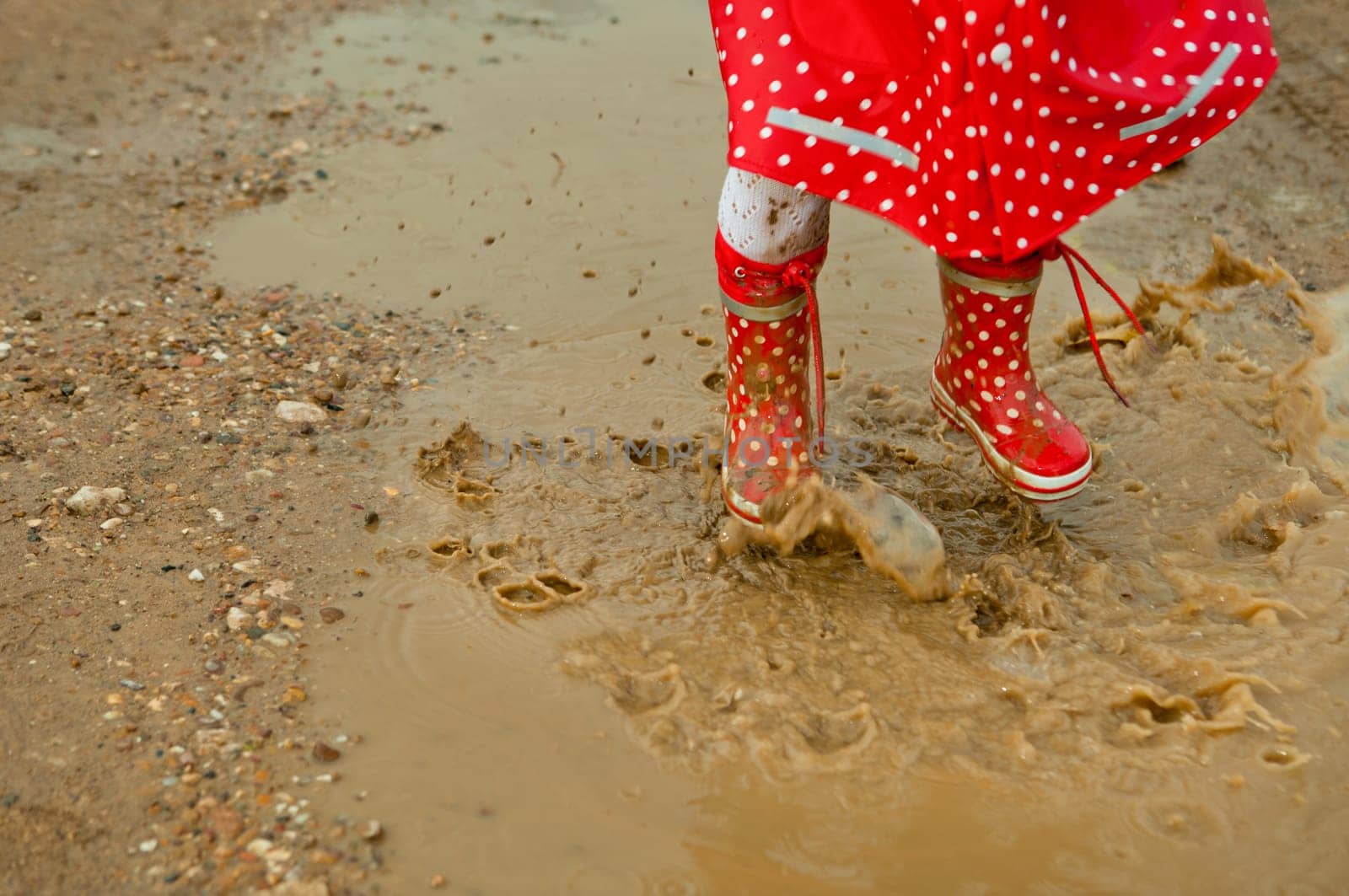 Detail of rainboots of a baby girl dressed with dotted raincoat in the puddles