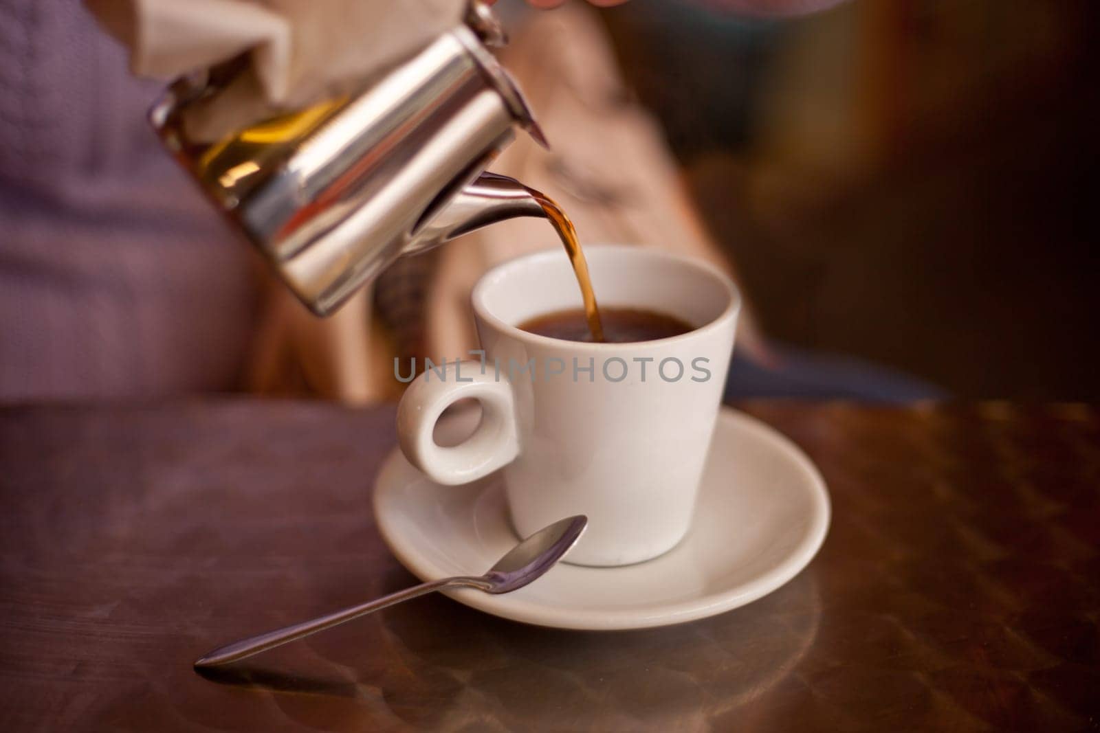 Serving tea from a metal teapont in a white ceramic cup on restaurant terrace table. Selective focus