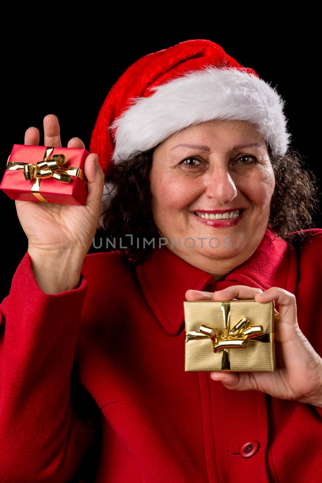Smiling elderly lady with red coat and Santa Claus hat is showing two small wrapped Christmas presents in her palms, one red, one golden. Isolated on black background. White teeth. Gift-giving theme..