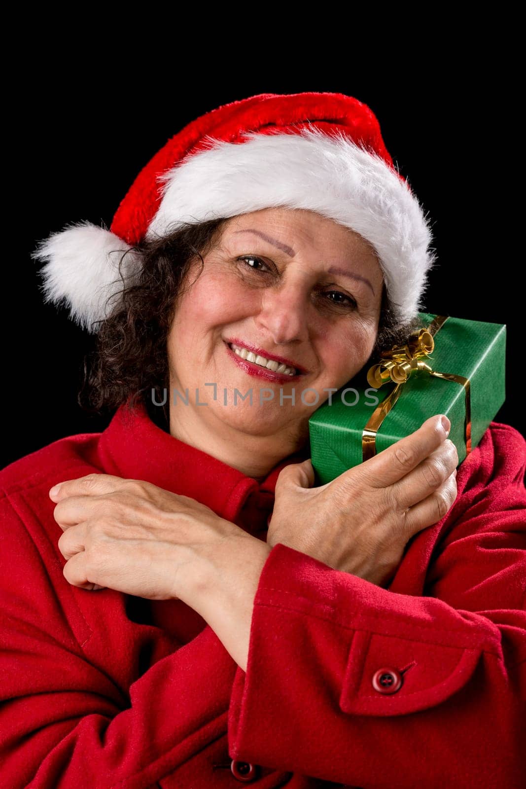 Smiling middle-aged lady is cuddling a green wrapped Christmas present. She is wearing a Santa Claus cap and a red coat. Vertical shot. Isolated on black background. Seasonal gift-giving theme..