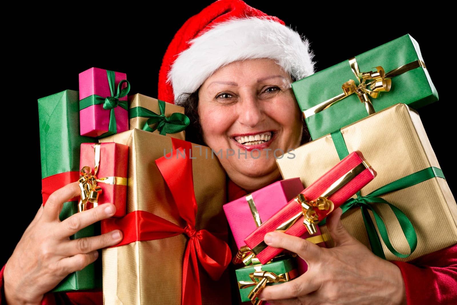 Honorable lady brimming over with joy is pressing ten wrapped Christmas presents to her chest. Santa Claus hat and coat. Smiling teeth. Isolated on black. December theme. Red, green and golden gifts..