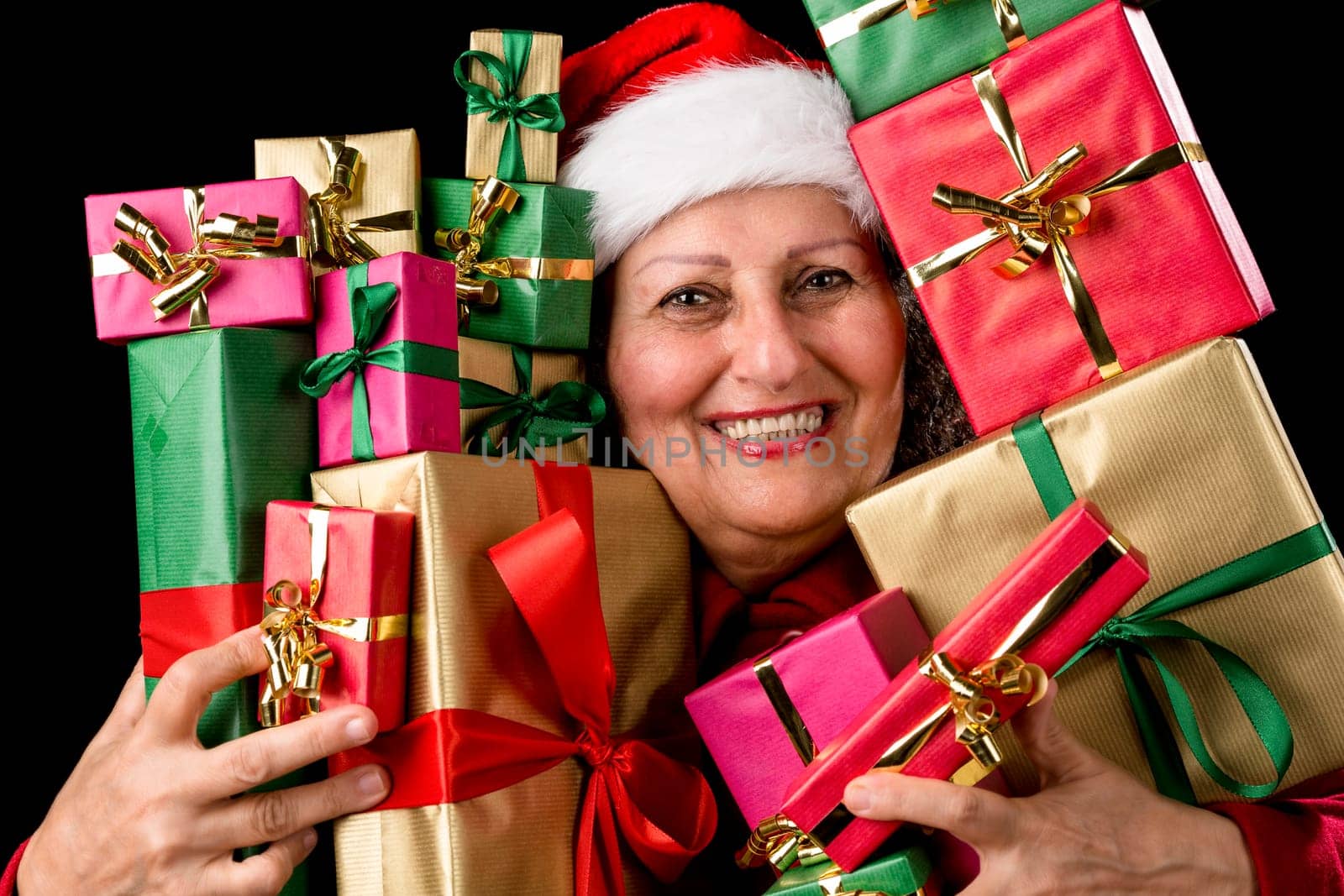 Cheerful elderly woman with Santa Claus hat is embosoming a host of Christmas presents. Santa Claus cap and white teeth. Gifts wrapped in plain red, gold and green. Isolated on black background..