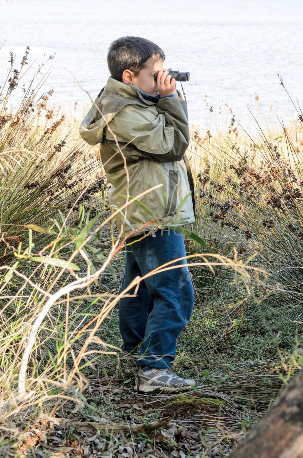 A school-aged boy looks through binoculars with a sea shore on the background.