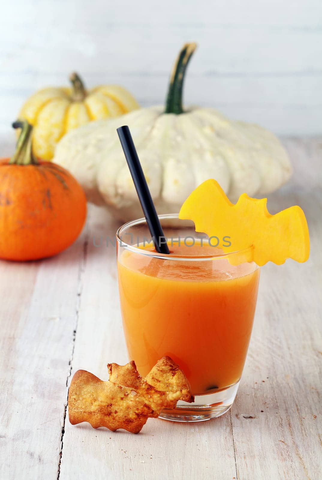 Glass of Halloween juice or vegetable smoothie served with a straw and scary bat shaped cookies and garnsih on rustic white wooden boards with pumpkins