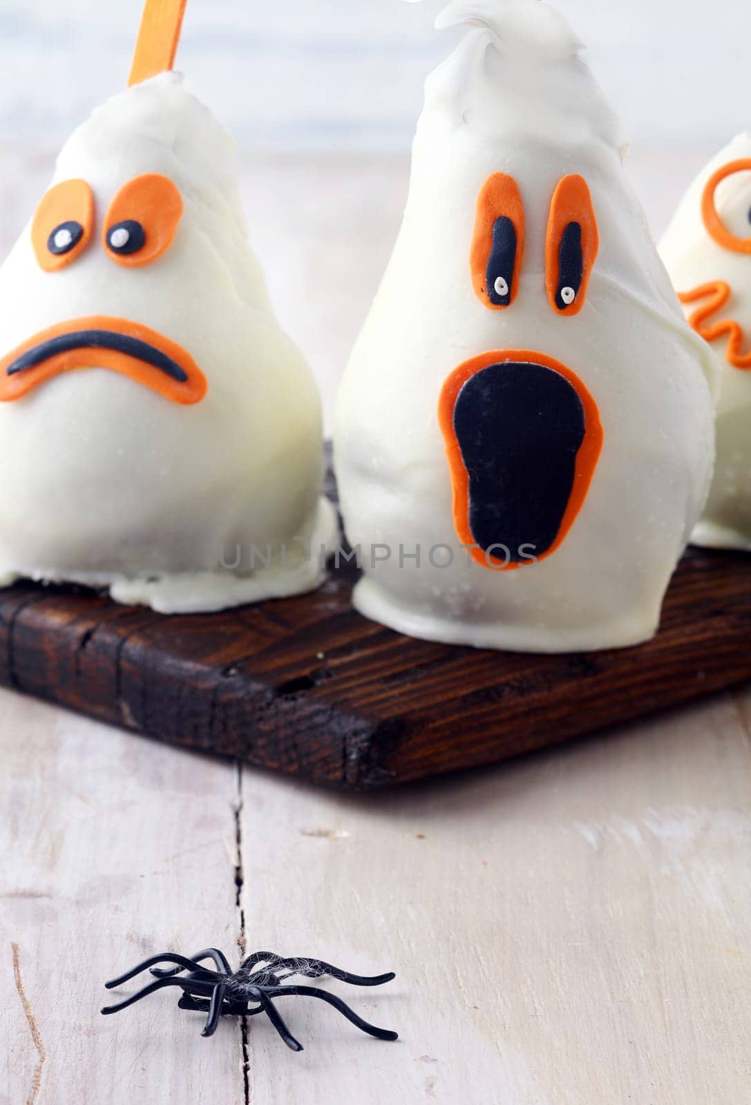 Scary edible Halloween party decorations with fresh pears covered in white chocolate and marzipan to resemble ghosts with spooky expressions on a rustic white table with a spider