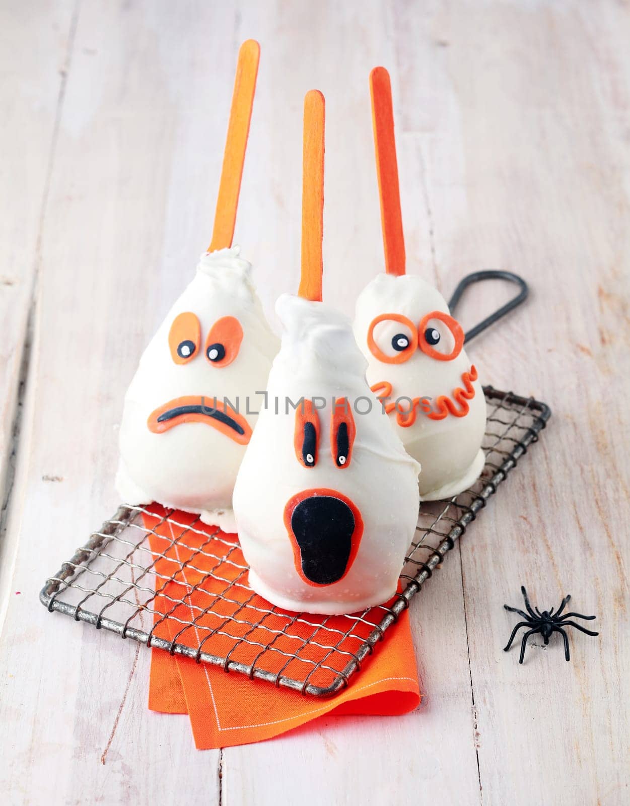 Ghostly edible trick-or-treat Halloween treats made from fresh pears coated in marzipan and chocolate with fun scary expressions with orange highlights cooling on a wire rack alongside a spider