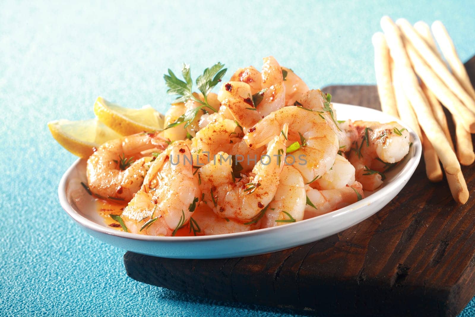 Healthy gourmet appetizer of grilled pink prawns or shrimp seasoned with olive oil and chopped fresh herbs and served with bread sticks on a blue textured table, low angle view