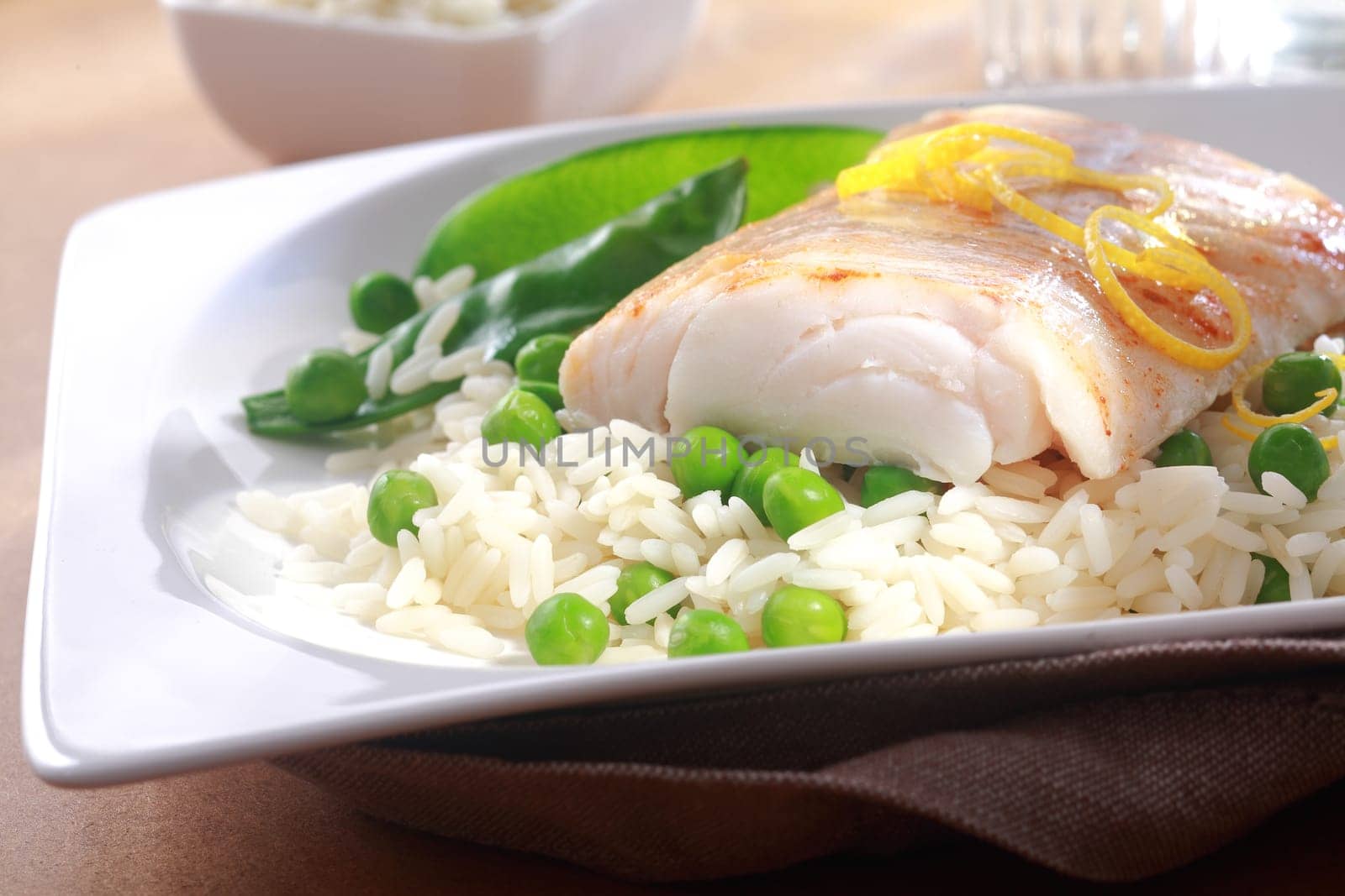 Healthy meal of baked fish fillet garnished with fresh lemon zest, rice and peas served on a white plate