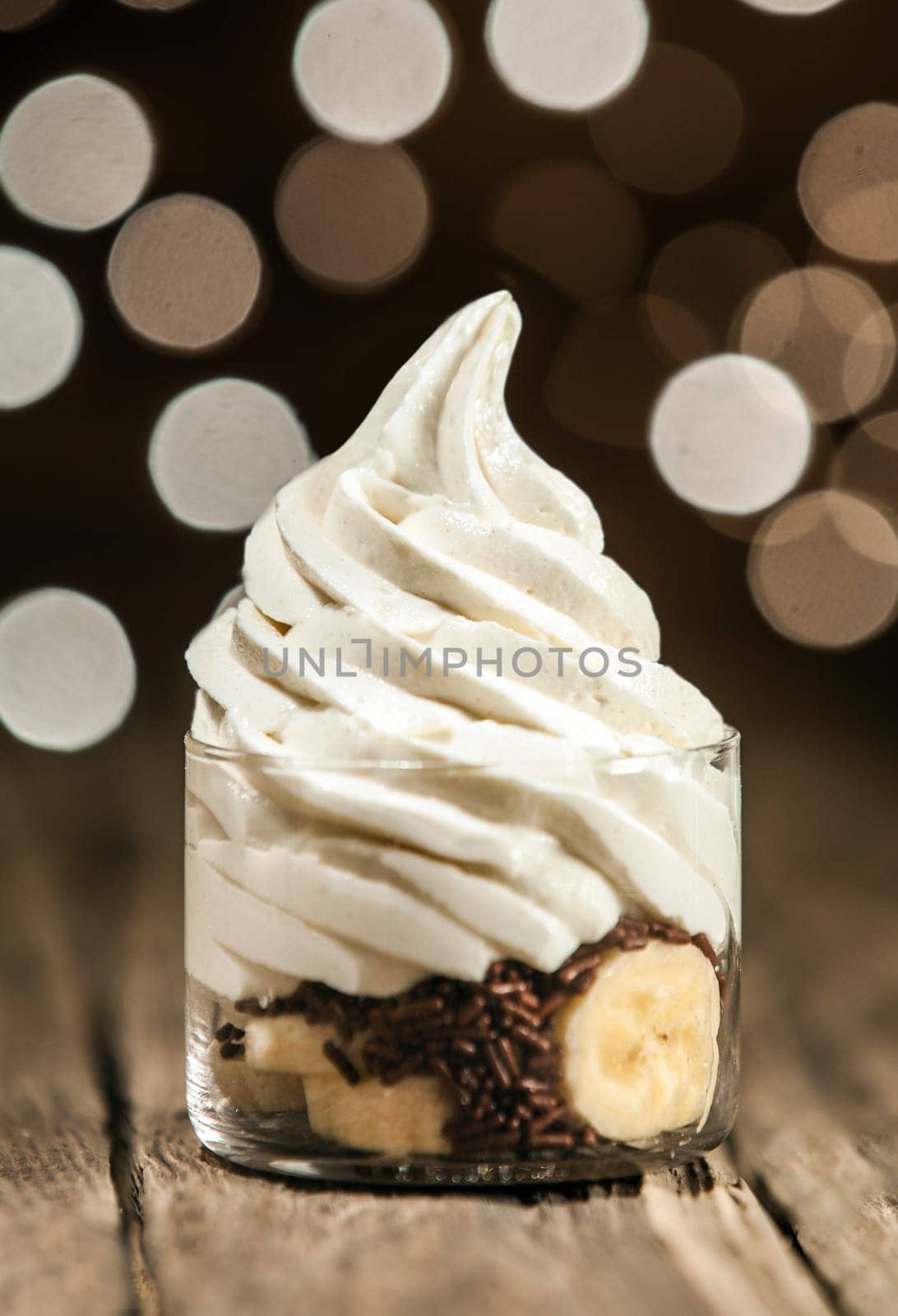 Delicious party dessert of sliced fresh banana and chocolate sprinkles topped with a swirl of low-fat frozen yogurt on a rustic wooden counter with a bokeh of sparkling lights