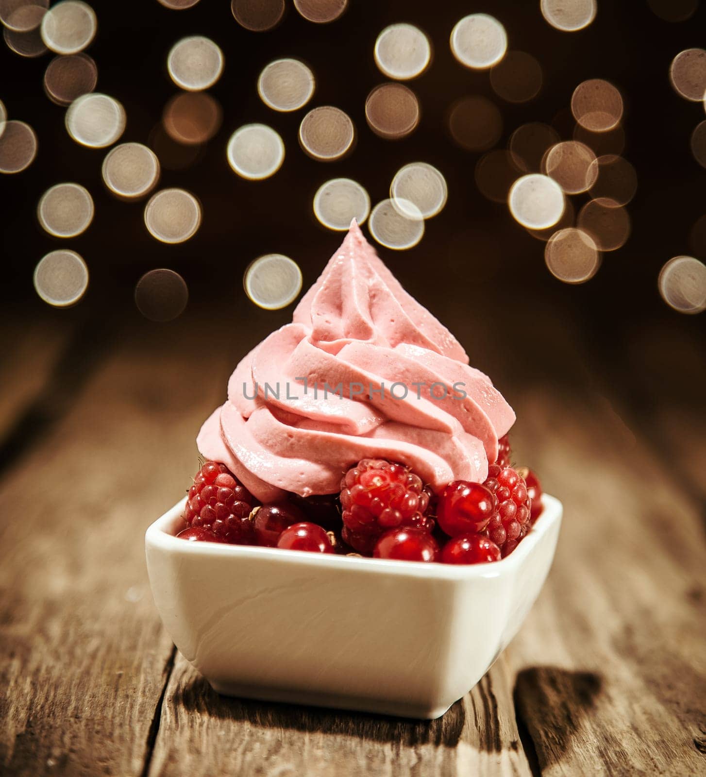 Fresh raspberries and redcurrants served in a dish topped with a swirl of frozen berry yogurt on a wooden bar counter against a bokeh of twinkling party lights