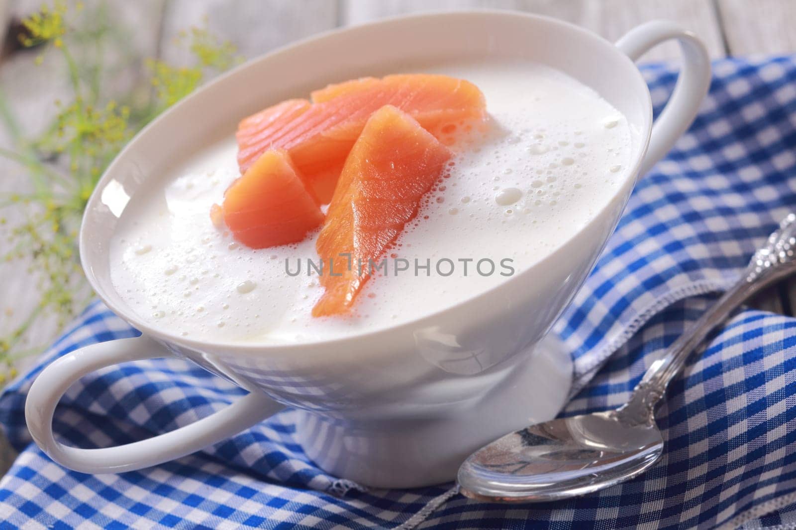 Bowl of gourmet chowder with raw salmon pieces in a frothy broth in an elegant white china bowl over a blue and white checked napkin