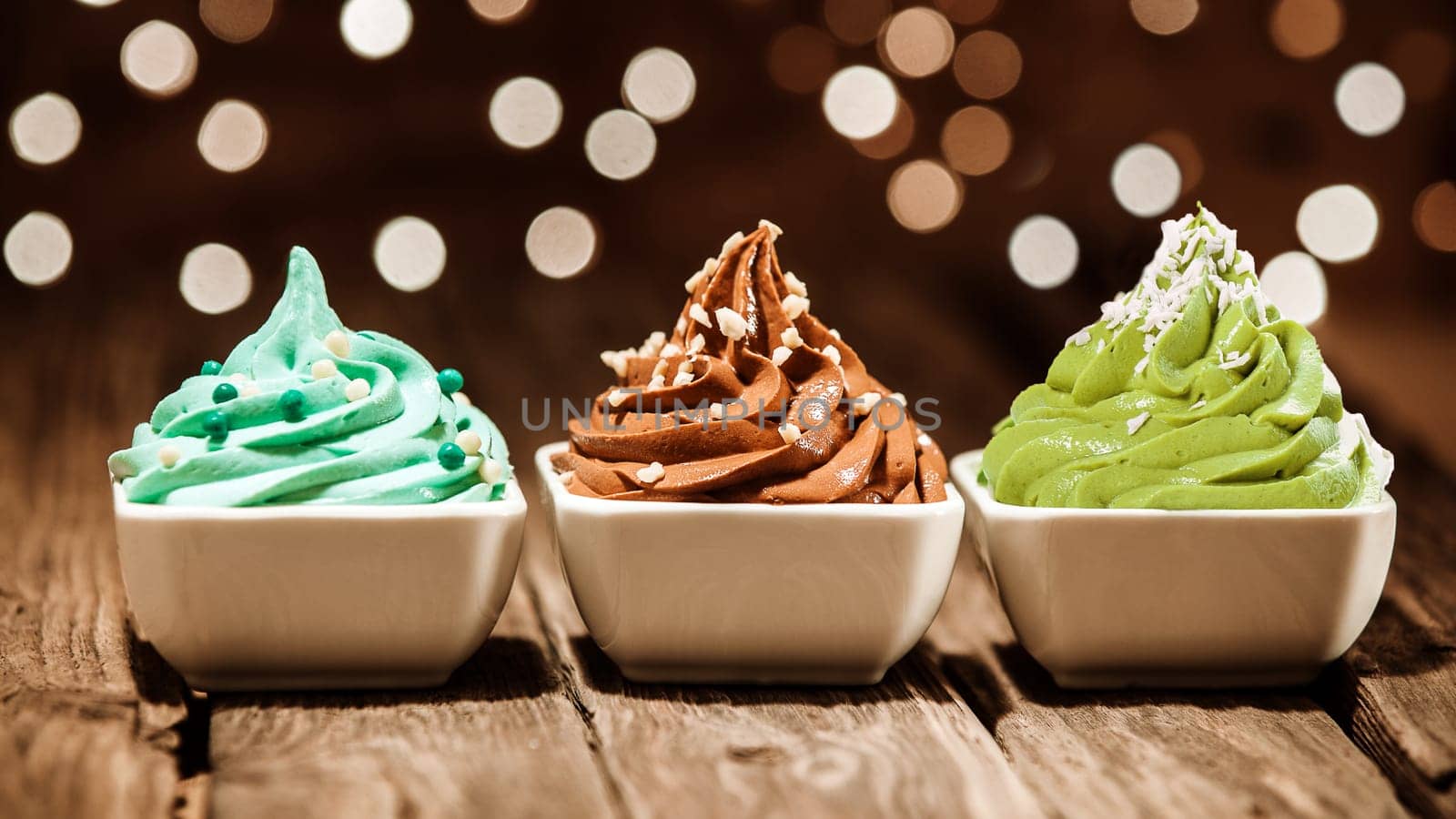 Colorful row of three different frozen yogurt desserts in blue, brown and green garnished with nuts and sugar pearls at a party with a sparkling background bokeh of lights