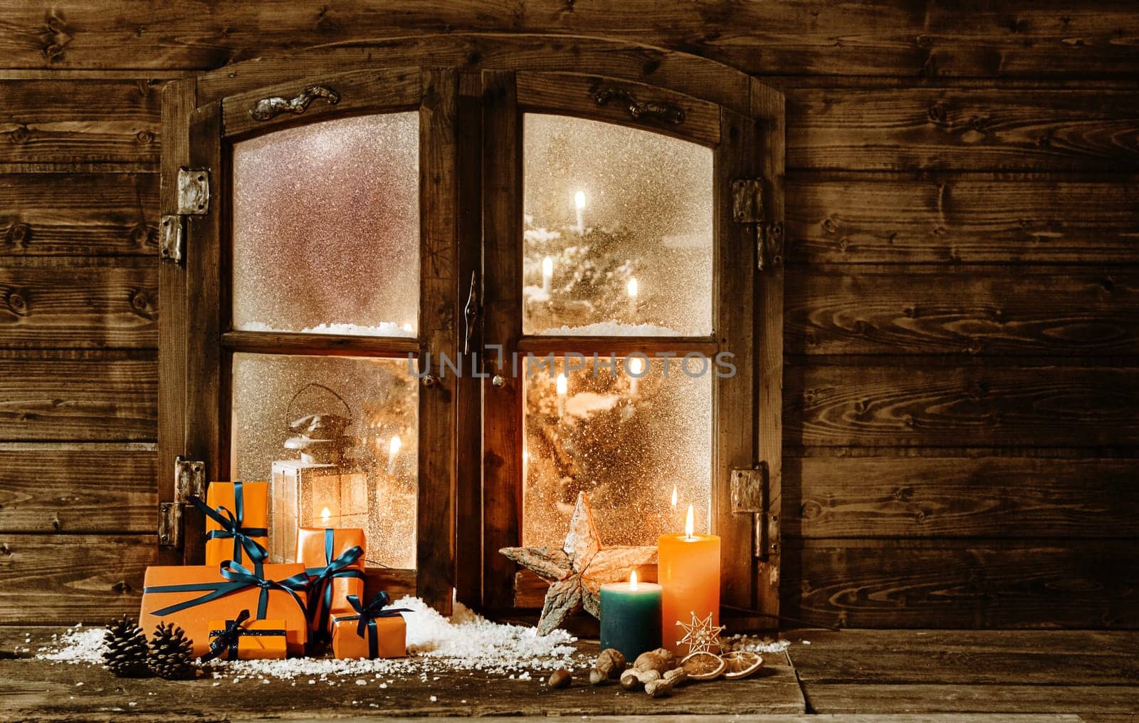 Festive wooden Christmas cabin window with gift-wrapped colorful orange presents, burning candles and decorations in winter snow and a glimpse of a decorated Christmas tree through the frosted window